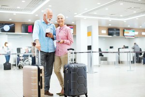 Closeup rear view of a cheerful senior couple looking at arrival departure board at an airport. They are standing in the middle of a brightly lit corridor, some blurry people in background.
