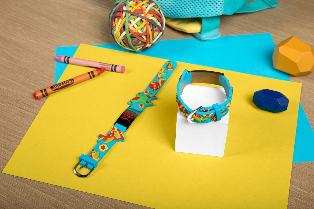 Two colorful MedicAlert QR code IDs with cartoon designs displayed on a yellow background, surrounded by crayons, a rubber band ball, and geometric shapes