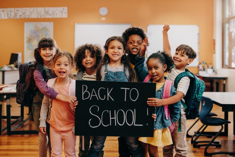 Group of cheerful children in a classroom holding a sign that reads 'Back to School!' with backpacks on their backs, smiling and excited.