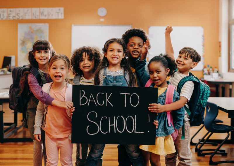 Group of cheerful children in a classroom holding a sign that reads 'Back to School!' with backpacks on their backs, smiling and excited.