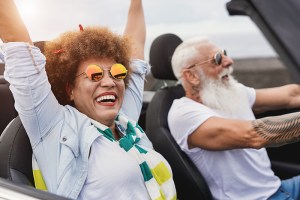 Trendy senior couple having fun inside convertible car - Multiracial mature people on a road trip with cabriolet car